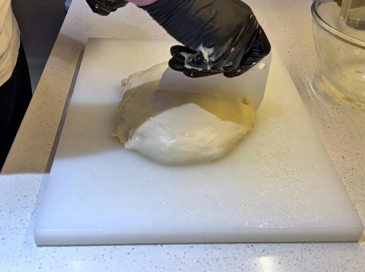 Manos con guantes negros utilizando un cortamasas de plástico blanco para partir un trozo grande de masa por la mitad.