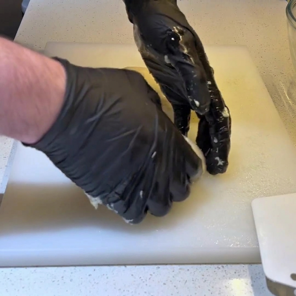 Manos enguantadas remetiendo los bordes de una porción de masa por debajo para formar una bola tensa y redonda.