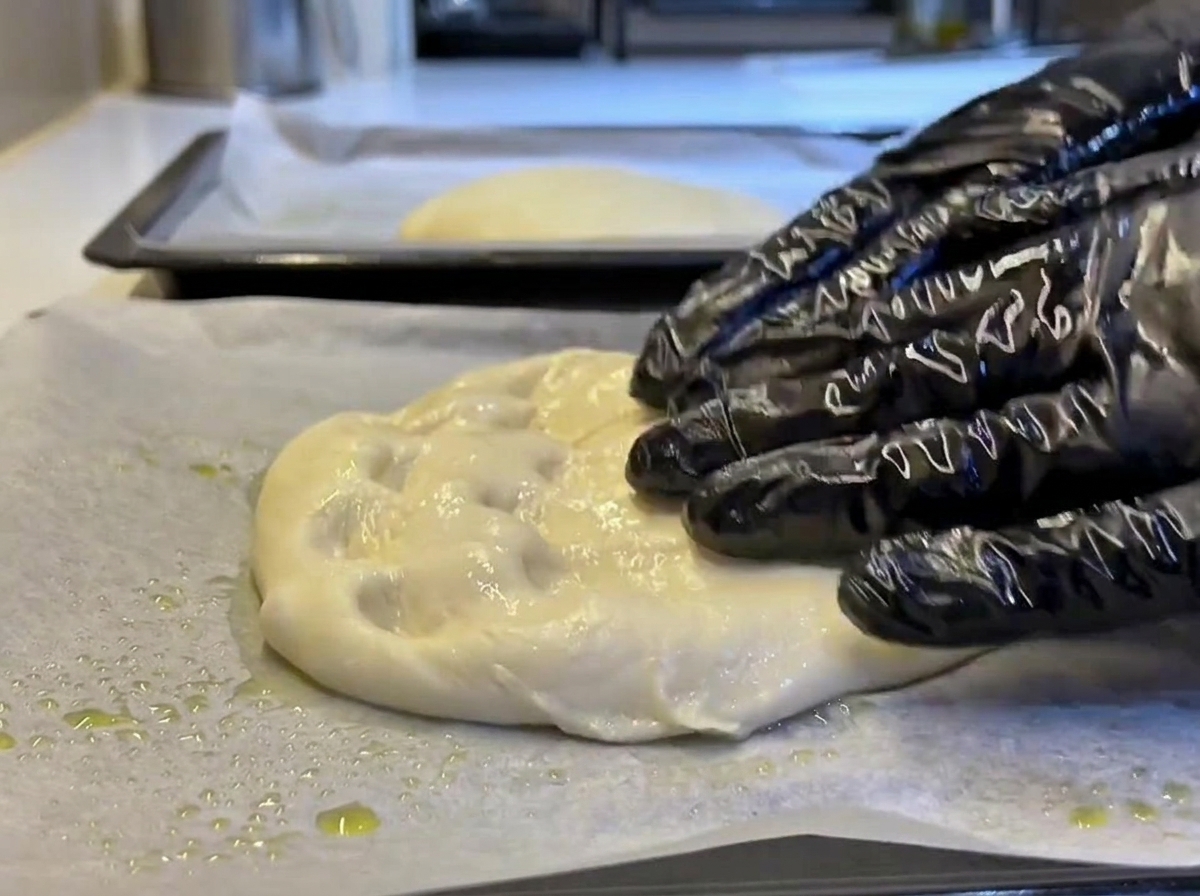 Manos enguantadas presionando y estirando suavemente un trozo de masa blanda hasta formar un rectángulo sobre papel de horno engrasado.