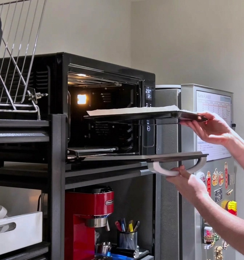 Una persona volviendo a meter en el horno caliente una pizza rectangular cargada de queso para su horneado final.