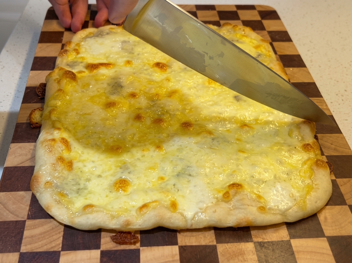 Un cuchillo de chef grande cortando una pizza rectangular de cuatro quesos recién horneada y burbujeante sobre una tabla de cortar de madera a cuadros.