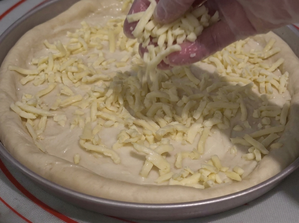 Una mano enguantada espolvoreando queso rallado sobre la masa con salsa.
