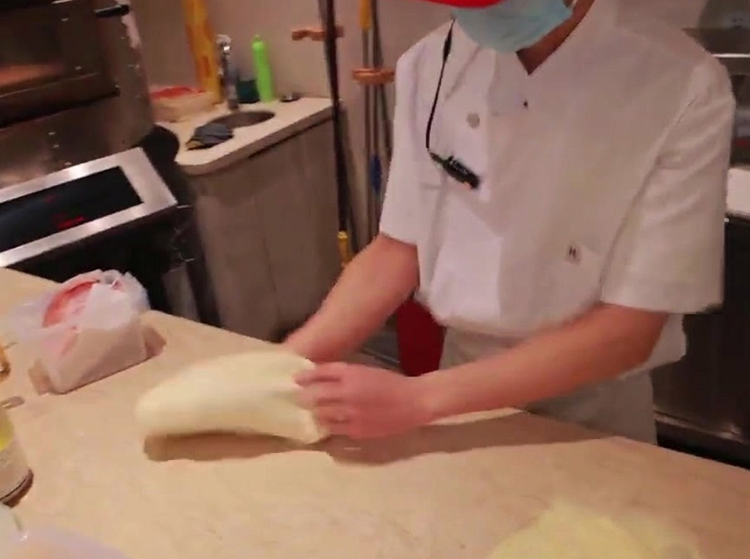 Manos de un chef estirando una bola de masa de pizza sobre una mesada de mármol enharinada.