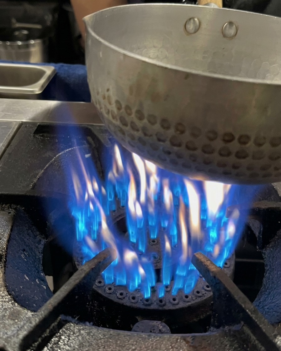 Caldo de hueso de cerdo siendo vertido en una pequeña olla sobre una llama de gas azul activa.