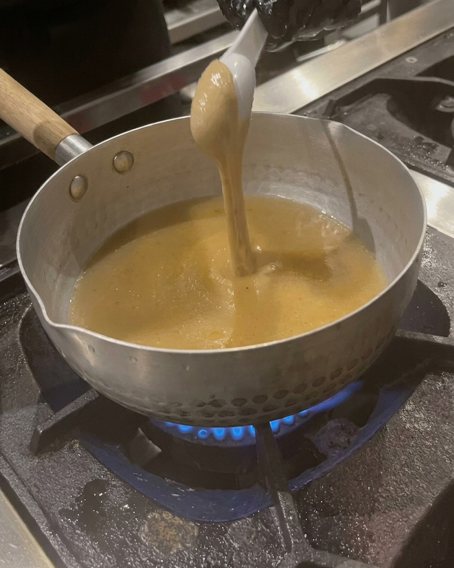 Una cucharada de pasta de sésamo espesa de color tostado siendo mezclada en el caldo hirviendo a fuego lento.