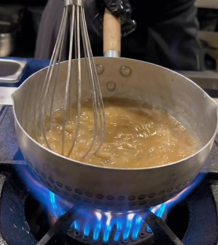 Fideos ramen siendo agitados con un batidor de alambre dentro de una olla de agua hirviendo.