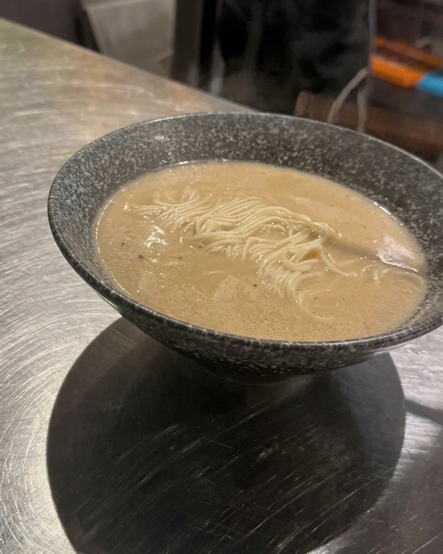 Una cucharada de carne picada sazonada siendo colocada en el centro de un tazón de fideos ramen con caldo.