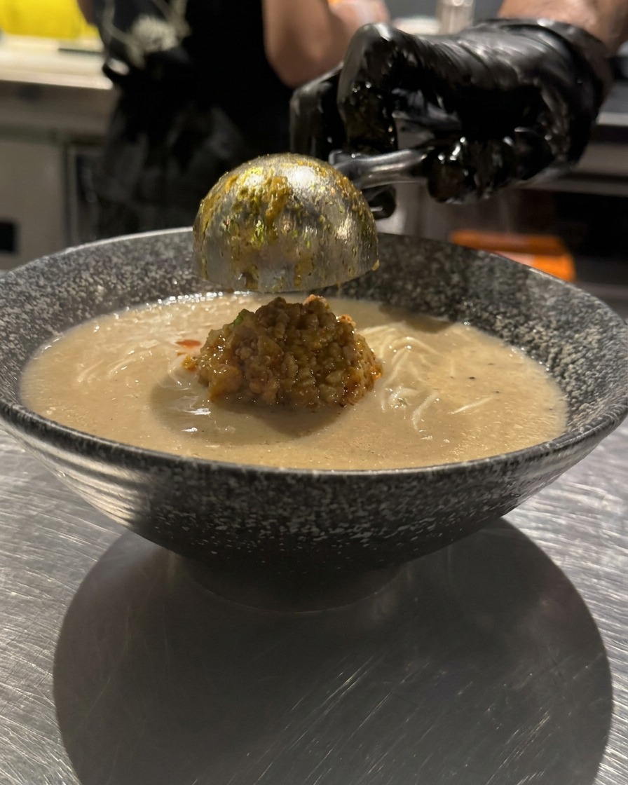 Una mano con guante rociando aceite de chile rojo brillante en patrones circulares sobre la superficie de un tazón de ramen.