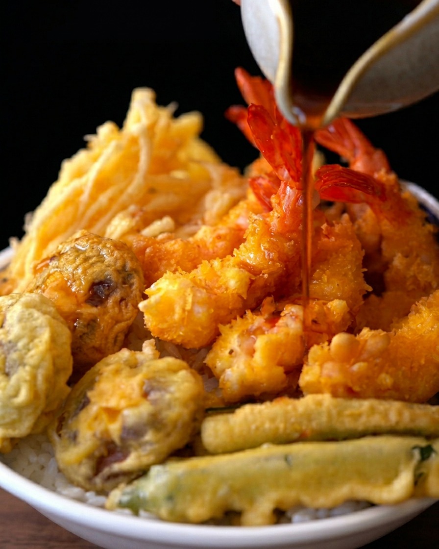 Una salsa oscura y sabrosa siendo vertida desde una jarra de cerámica sobre un tazón de arroz coronado con gambas y verduras en tempura.