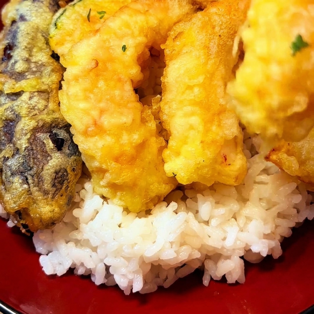 Piezas de tempura frita dorada siendo colocadas sobre una cama de arroz blanco en un tazón rojo.