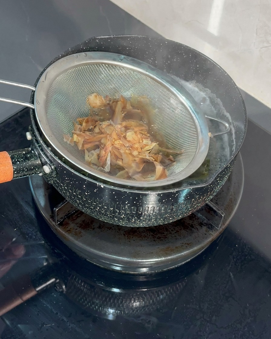 Un colador de metal de malla fina lleno de copos de bonito siendo sumergido en una olla de agua hirviendo.