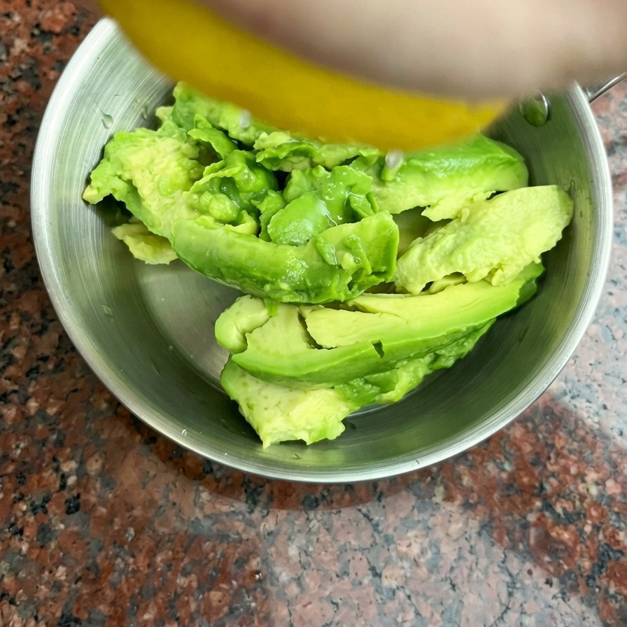 Jugo de limón fresco siendo exprimido a mano sobre mitades de aguacate dentro de un pequeño bol de acero inoxidable.