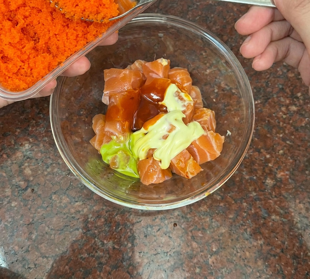 Una cuchara de plata añadiendo huevas de pescado de color naranja brillante a un bol con salmón en cubos.