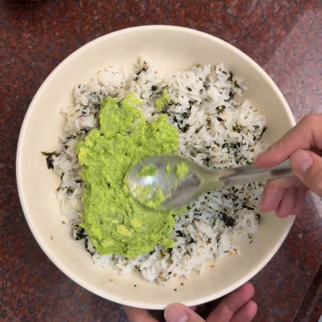 Una cuchara extendiendo puré de aguacate de color verde brillante sobre un bol de arroz blanco aderezado con tiras de alga oscura.