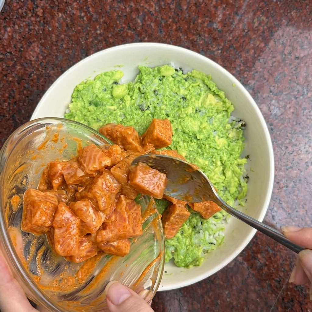 Una mano utilizando una cuchara para colocar cubos de salmón marinado de color naranja sobre un bol de arroz y aguacate.
