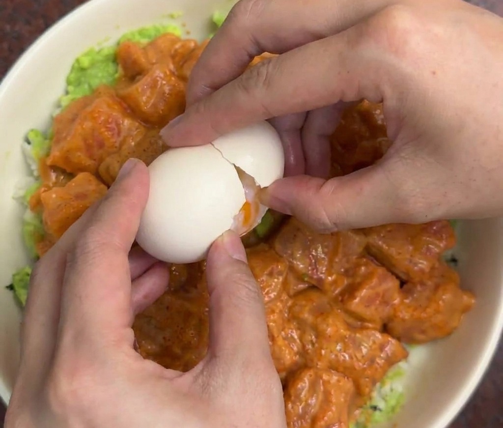 Manos cascando con cuidado un huevo onsen blanco en el centro de un bol de arroz con salmón y aguacate.