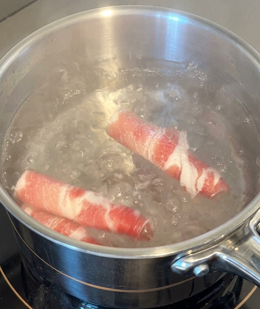 Dos láminas de ternera grasa cruda sumergiéndose en una olla de acero inoxidable con agua hirviendo.
