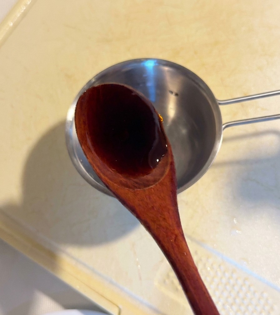 Una cuchara de madera vertiendo salsa de ostras oscura en un pequeño cuenco metálico con los ingredientes de la salsa.