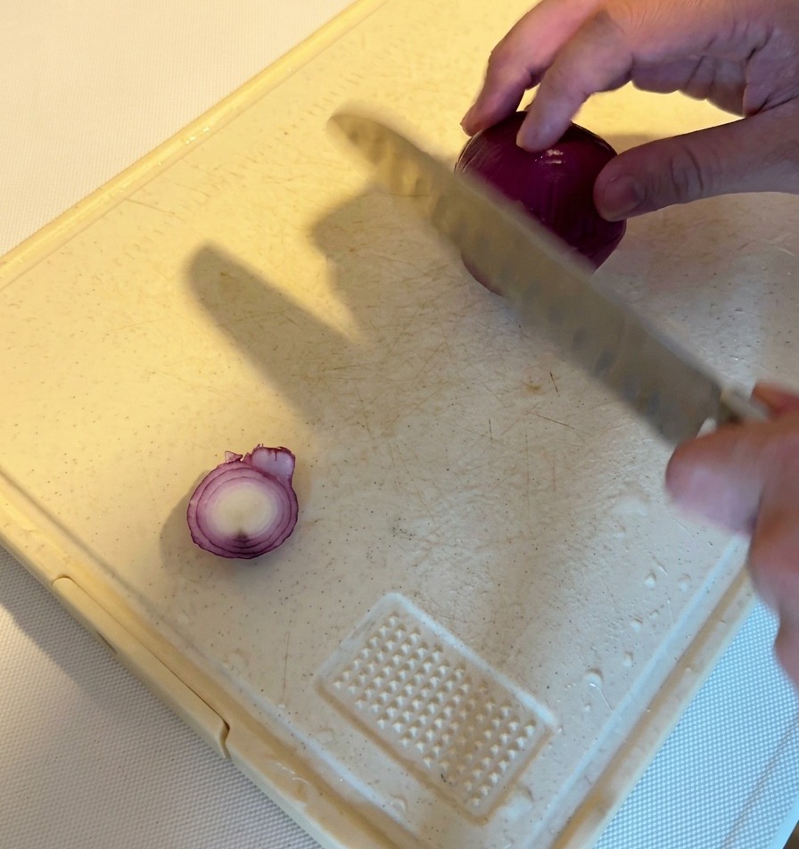 Una persona usando un cuchillo de chef para cortar una cebolla morada entera en juliana fina sobre una tabla de cortar blanca.
