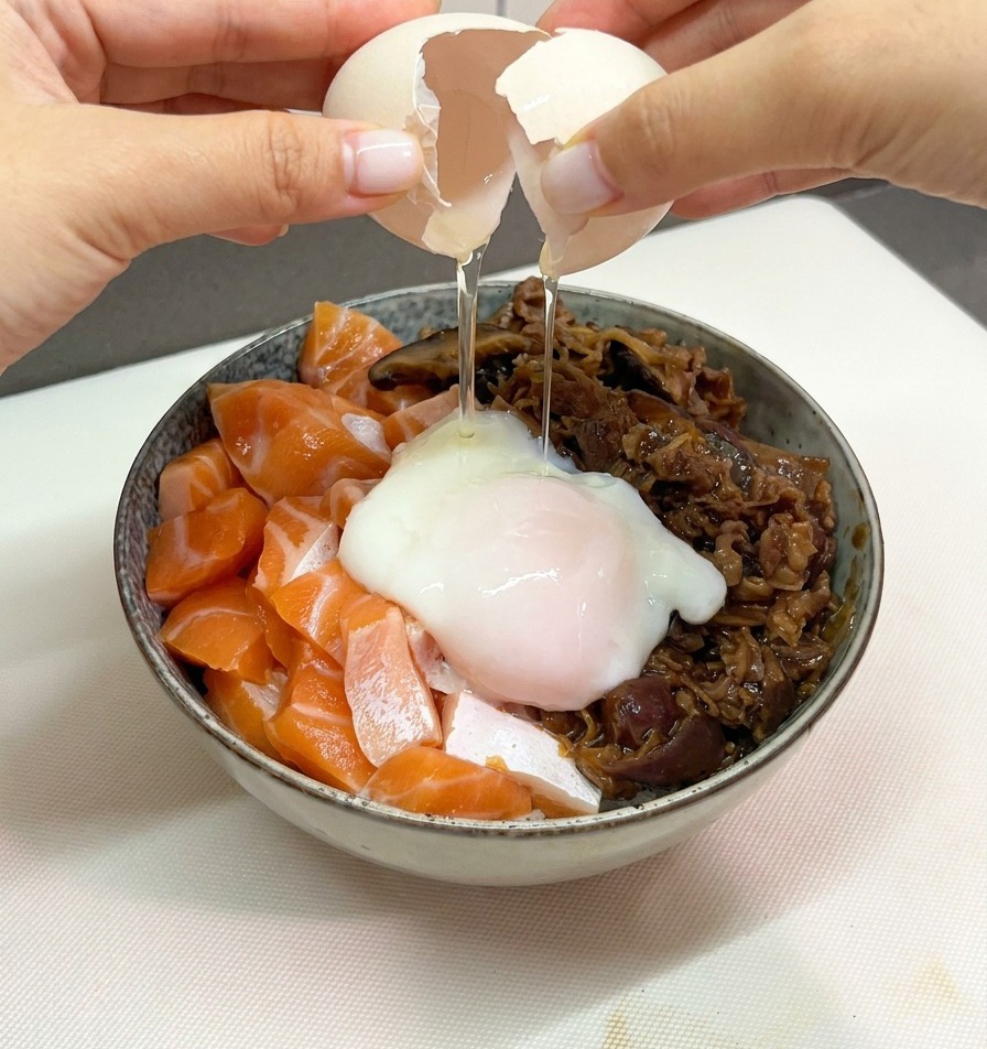 Dos manos abriendo la cáscara de un huevo para dejar caer un huevo onsen muy tierno sobre el tazón de arroz con salmón y ternera.