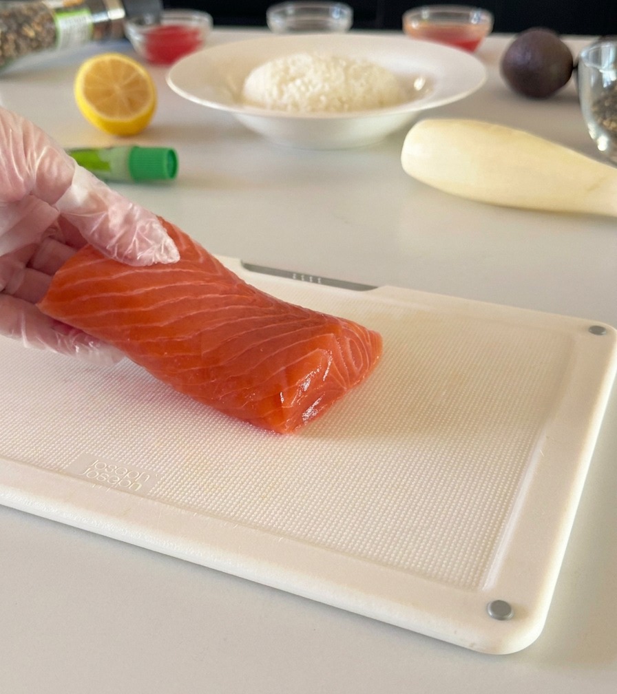 Una mano con guante sosteniendo un filete de salmón fresco sobre una tabla de cortar blanca, lista para trocearlo en dados.