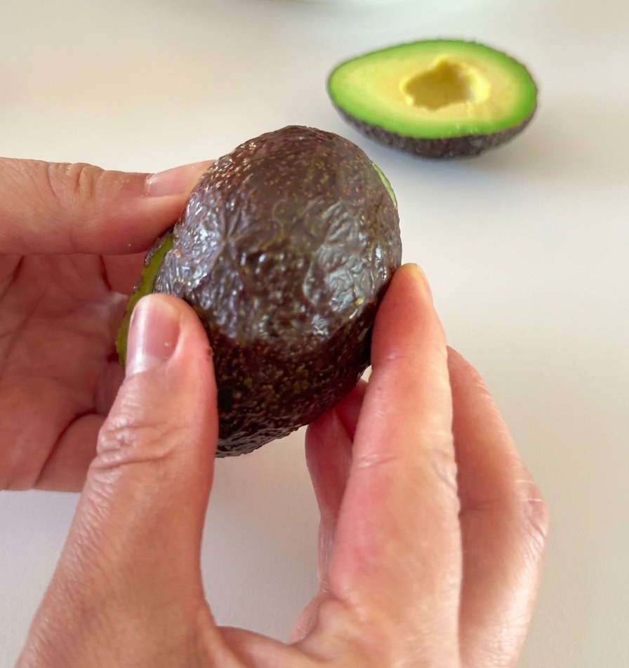 Dos manos retirando con cuidado la piel oscura de la mitad de un aguacate maduro.