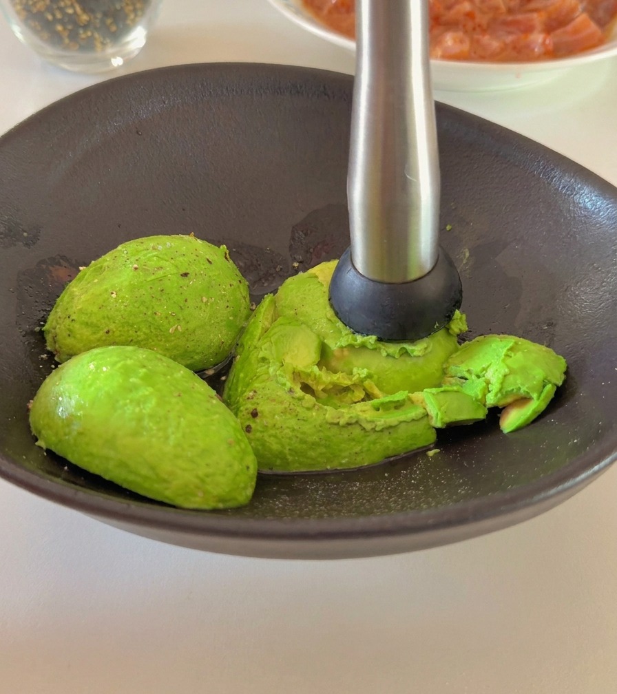 Una persona utilizando un pisapapas de metal para triturar mitades de aguacate en un cuenco de cerámica oscuro.