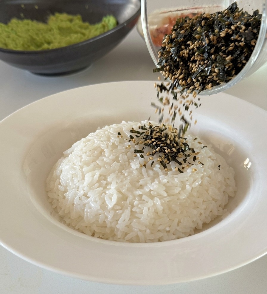 Una mezcla de tiras de alga negra y semillas de sésamo blanco cayendo desde un vaso de cristal sobre una base compacta de arroz blanco.