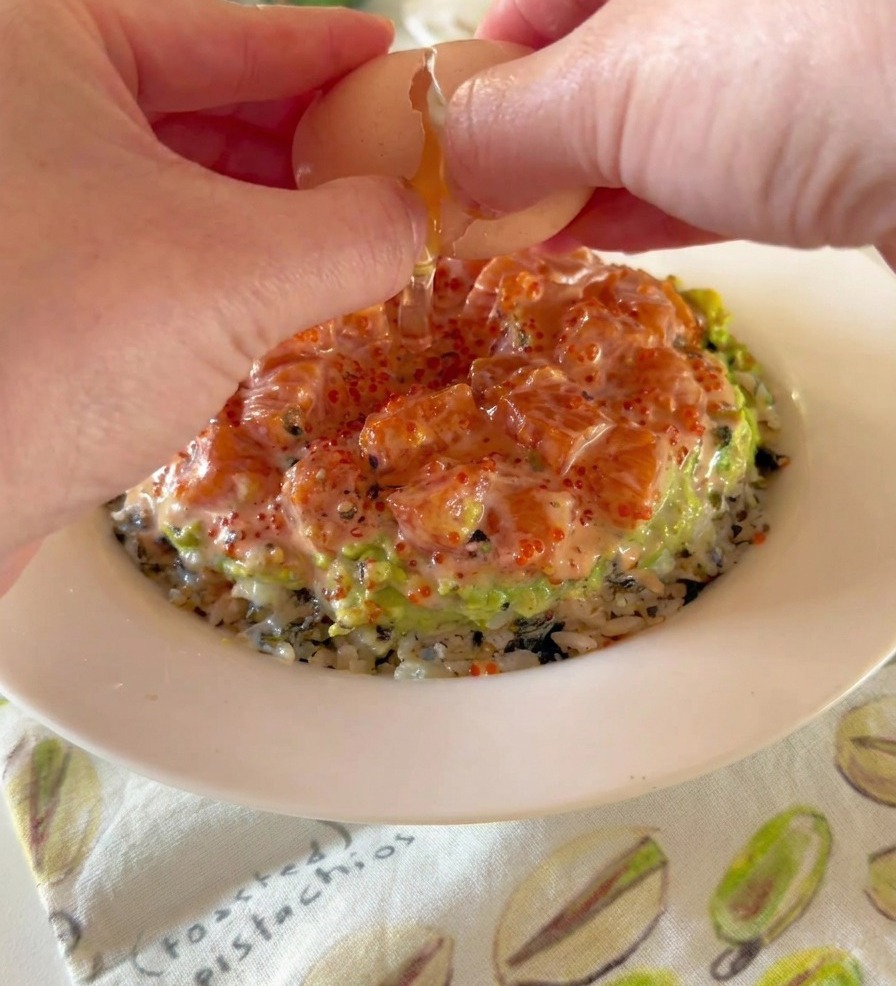 Unas manos cascando un huevo onsen de yema fluida sobre un tazón con capas de arroz, aguacate y salmón.