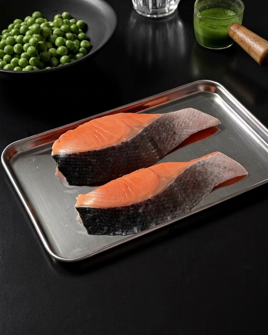 Dos filetes de salmón crudos en una bandeja de acero inoxidable, ligeramente sazonados con sal y pimienta negra.