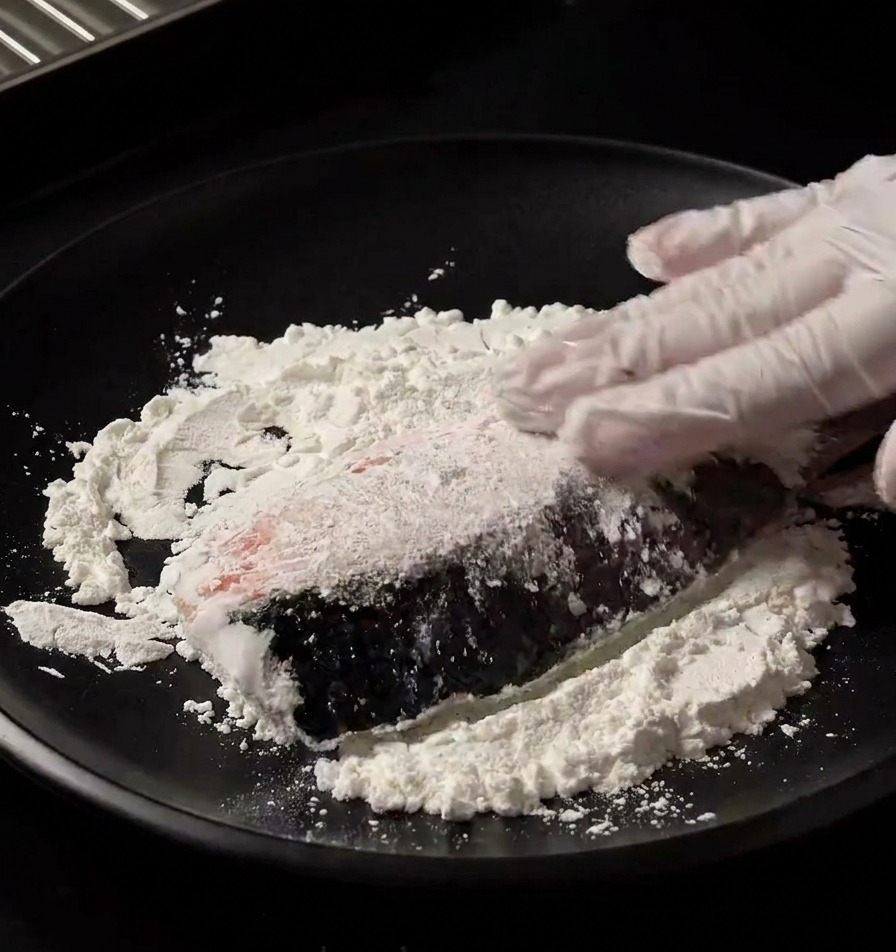 Una mano con guante presionando un filete de salmón fresco sobre un plato negro con fécula blanca para cubrirlo de manera uniforme.