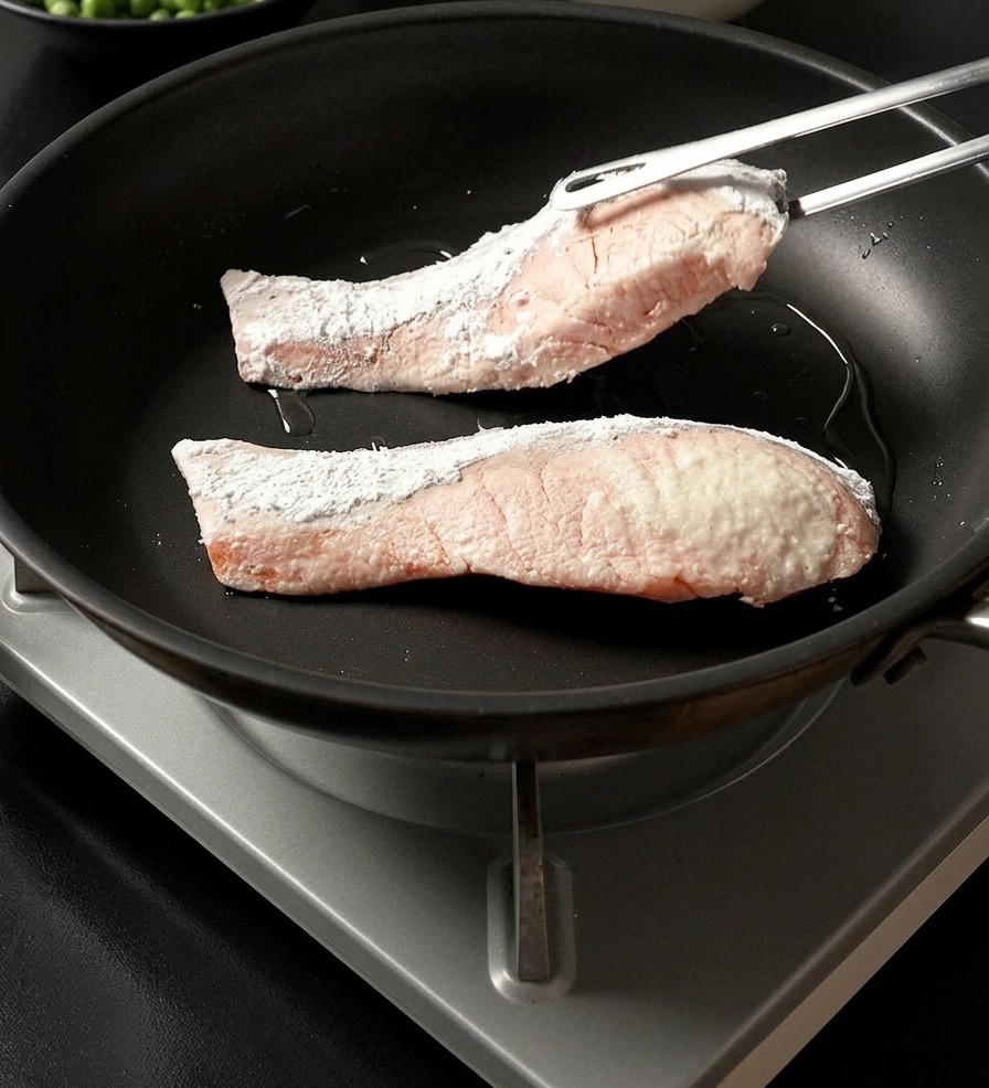 Unas pinzas metálicas colocando un filete de salmón rebozado en fécula dentro de una sartén caliente junto a otro filete.