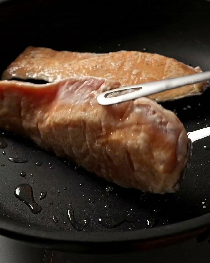 Primer plano de un filete de salmón al que se le da la vuelta en la sartén, mostrando su superficie dorada y crujiente.