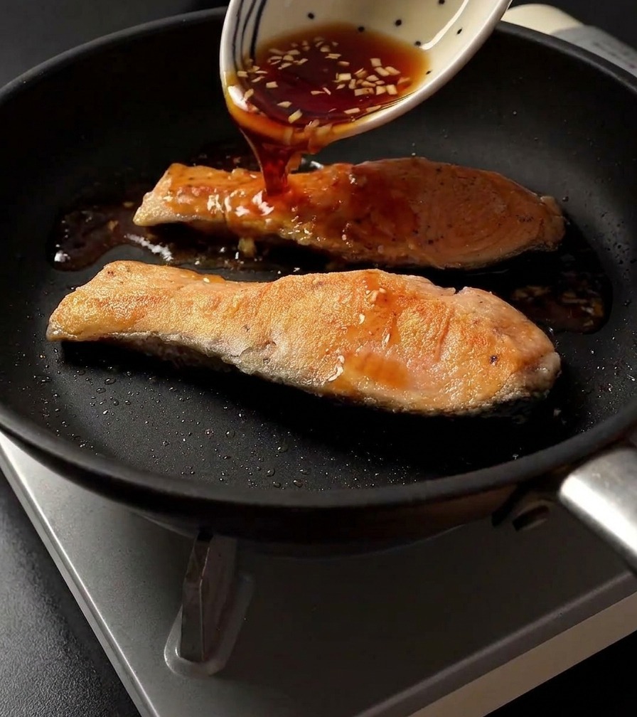 Vertiendo salsa teriyaki oscura desde un cuenco blanco decorado sobre dos filetes de salmón sellados en una sartén.