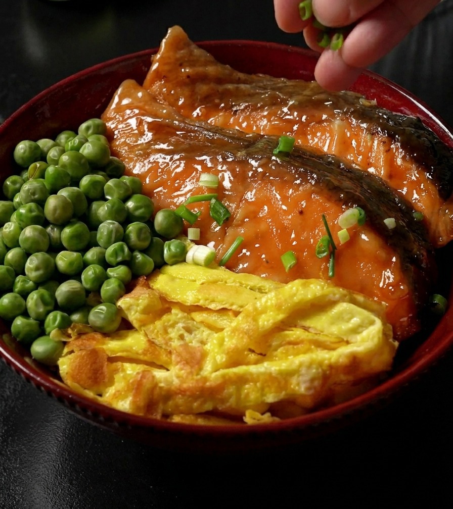 Una mano espolvoreando cebollino picado sobre un tazón de arroz terminado, coronado con salmón teriyaki, tiras de huevo y guisantes.