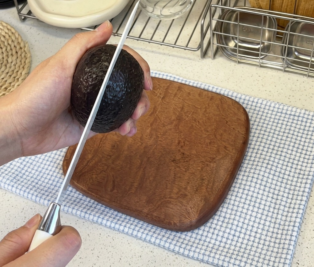 Manos de una persona sosteniendo un aguacate de piel oscura mientras lo cortan verticalmente con un cuchillo afilado de cocina.