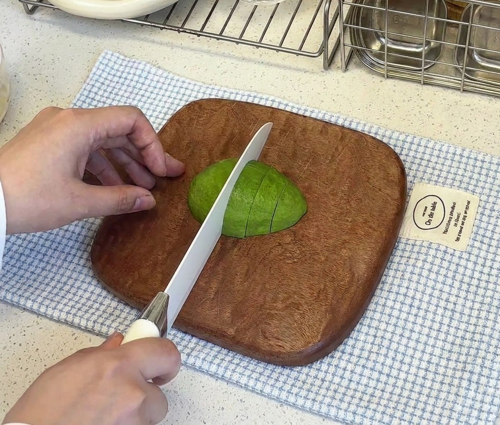 Una mano sujetando la mitad de un aguacate pelado sobre una tabla de cortar mientras un cuchillo blanco lo corta en cubos pequeños.