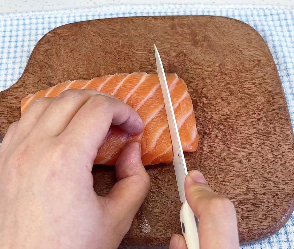 Primer plano de un cuchillo afilado cortando un filete de salmón fresco y marmoleado sobre una tabla de cortar de madera oscura.