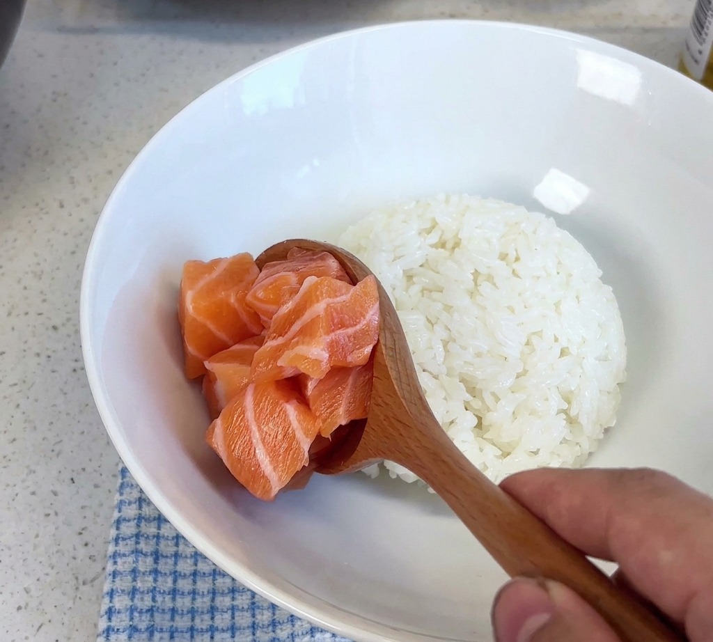 Una cuchara de madera levantando un montón de cubos de salmón fresco y colocándolos en un tazón blanco con arroz cocido.