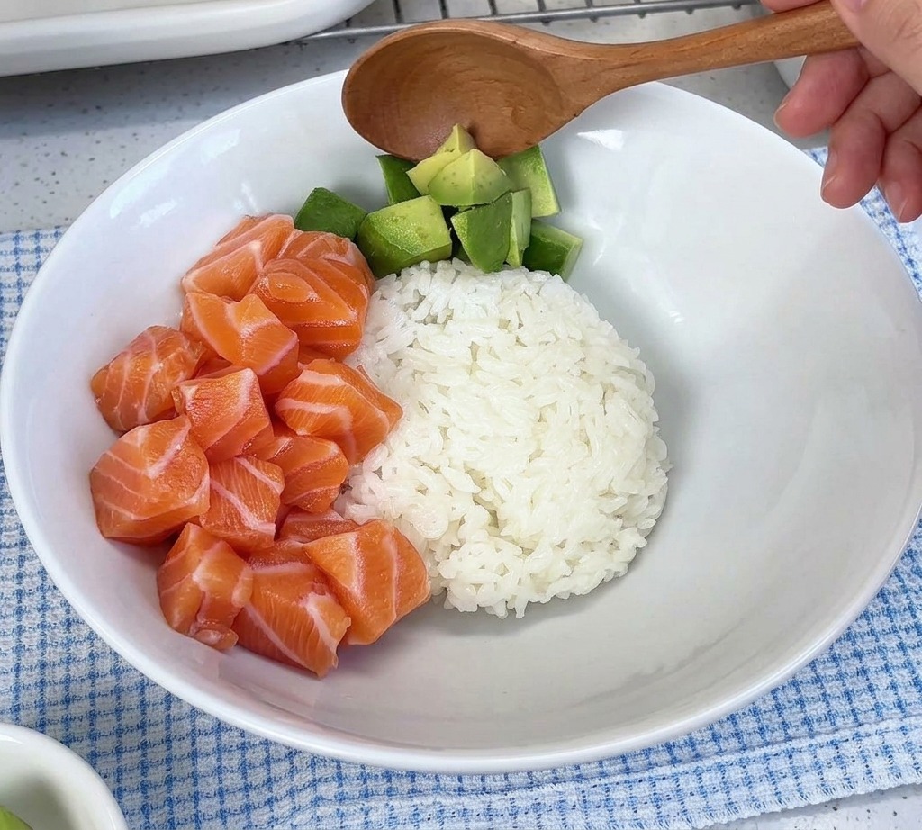 Aguacate en cubos siendo servido con una cuchara en un tazón blanco junto a un montón de cubos de salmón crudo sobre arroz.