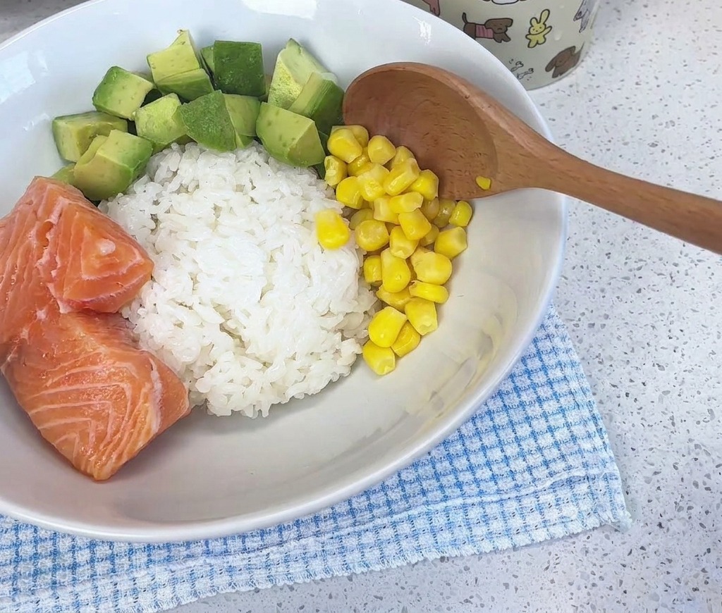 Una cuchara de madera agregando granos de maíz de color amarillo brillante en un tazón blanco lleno de arroz, aguacate en dados y trozos de salmón fresco.
