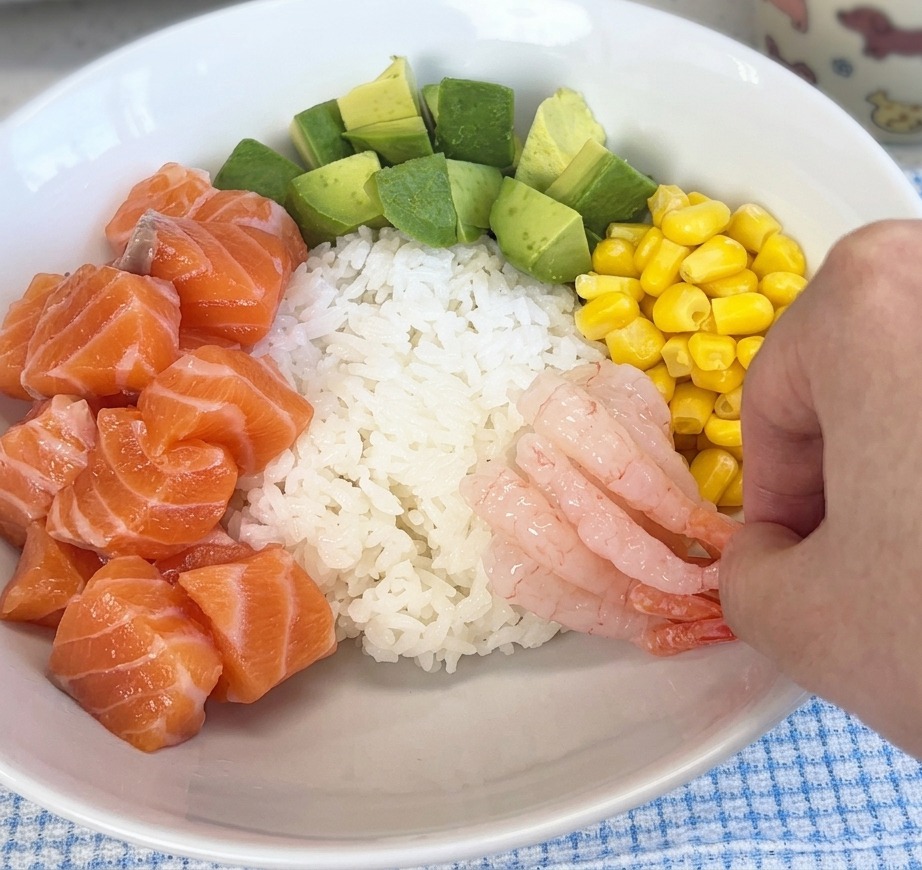 Una mano colocando camarones dulces, rosados y translúcidos sobre el arroz en un tazón junto con salmón y maíz.
