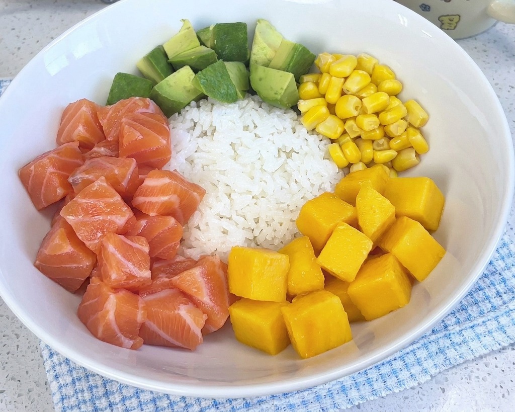 Un colorido poke bowl con arroz blanco, salmón, aguacate, maíz y camarones, que ahora incluye un montón de cubos de mango de color amarillo brillante.