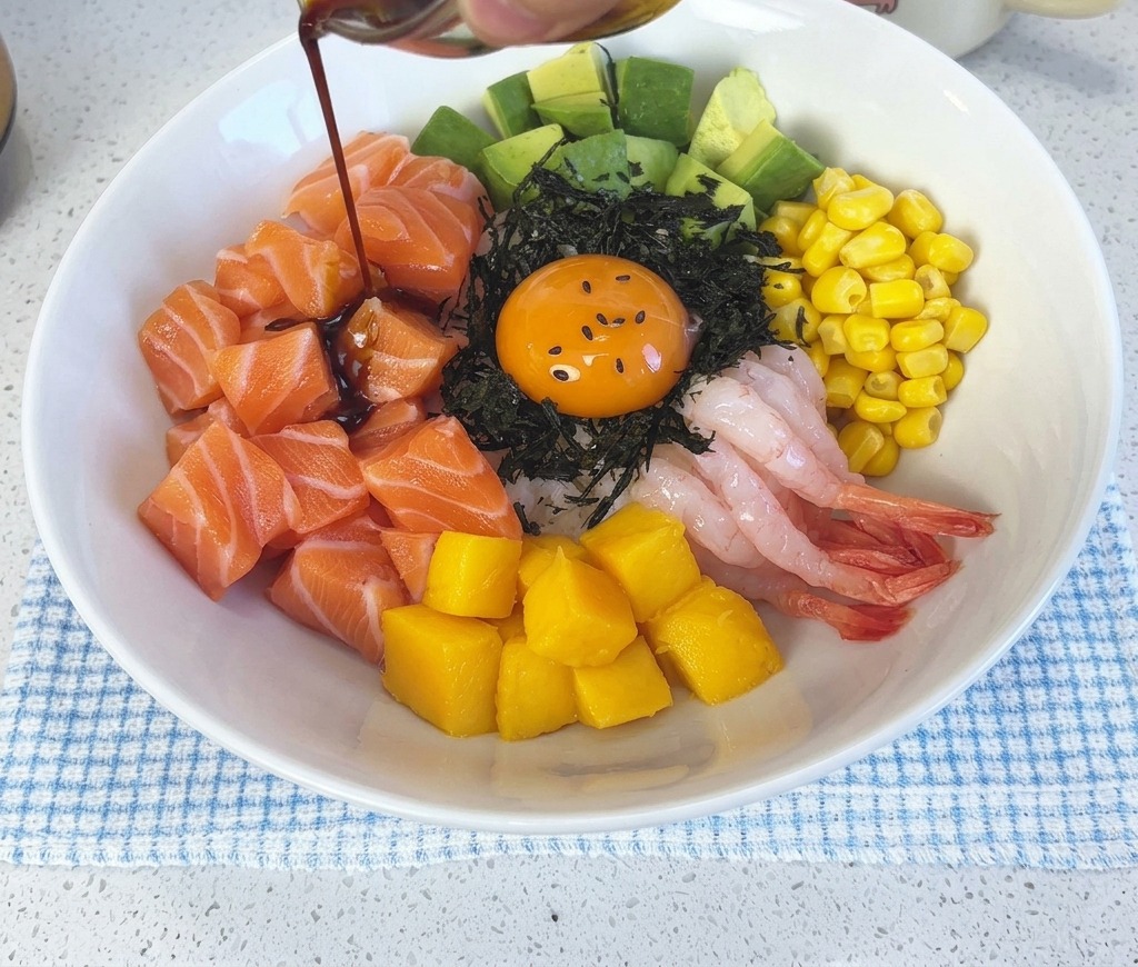Salsa oscura de soja y wasabi cayendo desde un pequeño tazón de vidrio sobre el poke bowl de salmón montado y la yema de huevo.