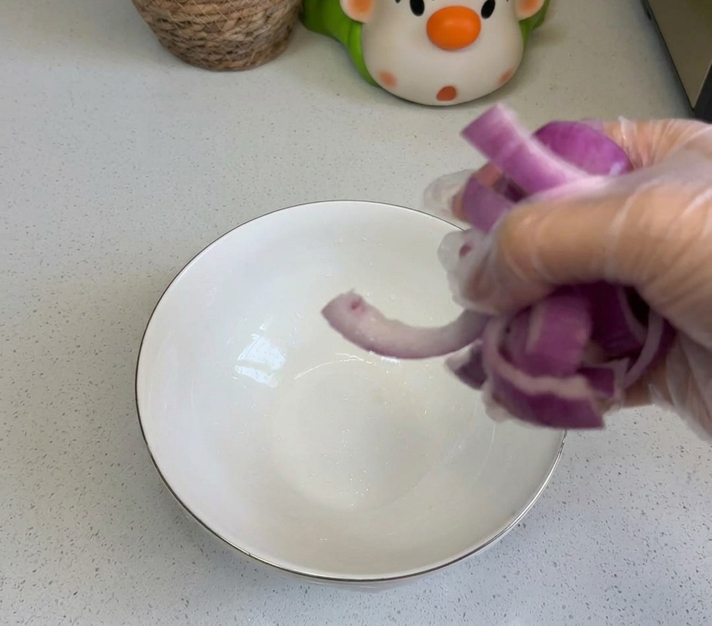 Una persona añadiendo cebolla morada cortada en un cuenco de cerámica blanca sobre la encimera de la cocina.