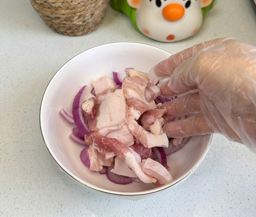 Lonchas de panceta de cerdo cruda introduciéndose en un cuenco blanco que ya contiene cebolla morada cortada.