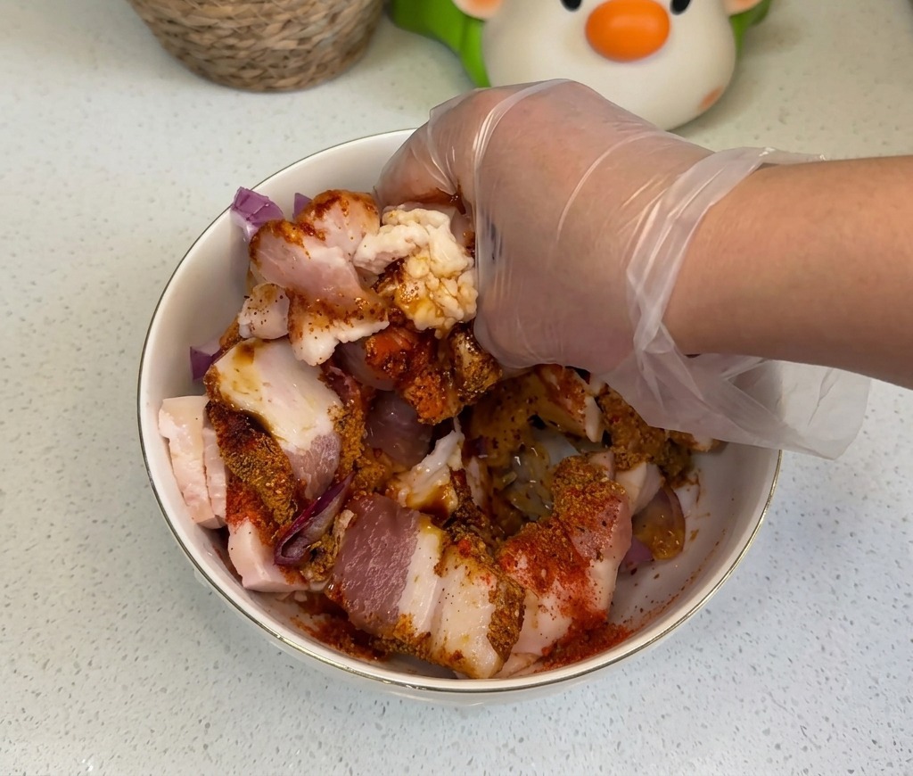 Una mano con un guante de plástico transparente mezclando bien las lonchas de panceta con cebolla y condimentos de colores.