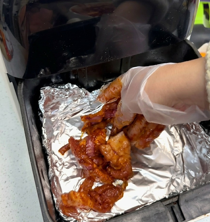 Una mano con guante transparente colocando lonchas de panceta marinada sobre papel de aluminio dentro de la cesta de la freidora.