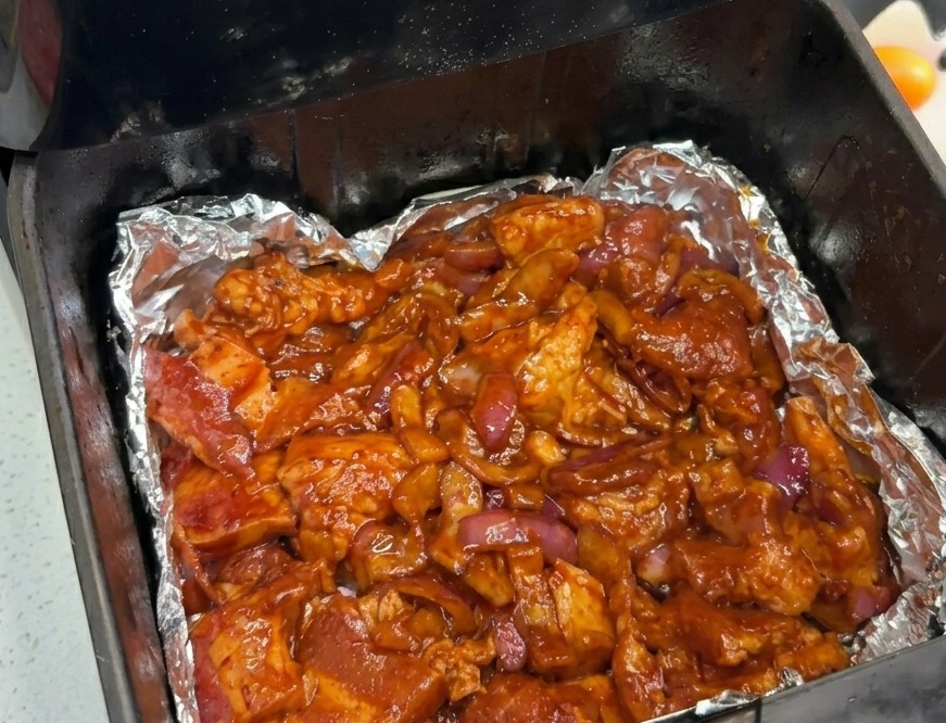 Panceta de cerdo marinada y cebolla morada distribuidas uniformemente en el fondo de una freidora de aire con papel de aluminio.