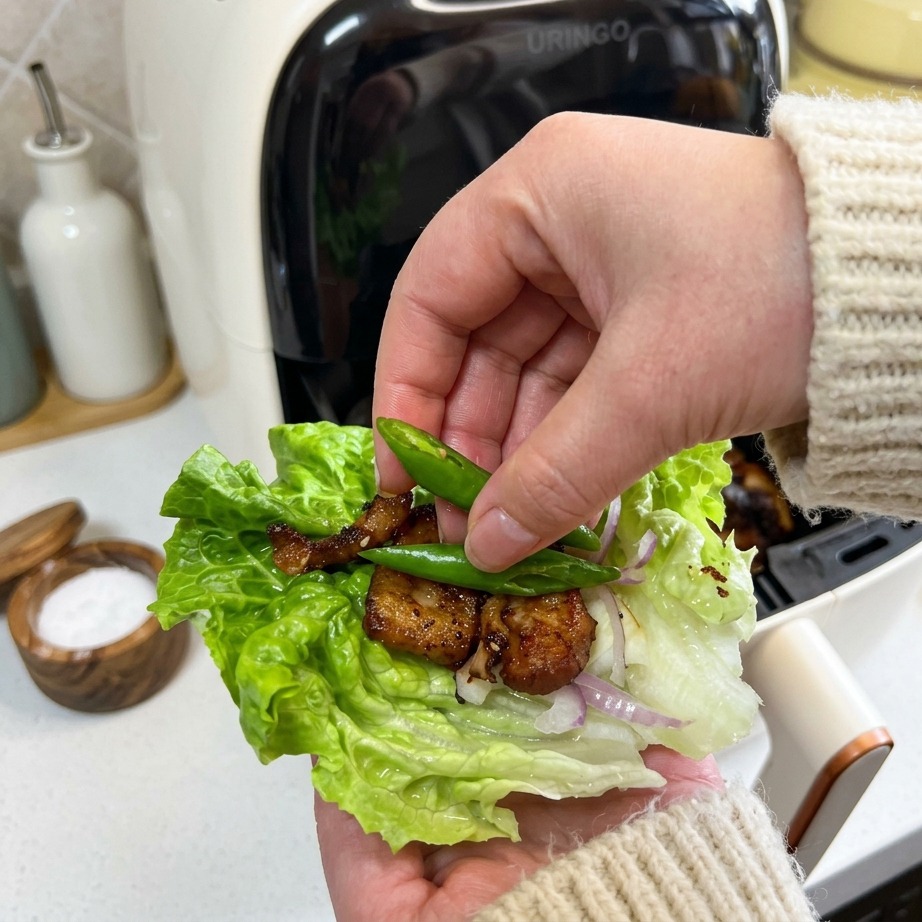 Una mano colocando dos pequeños chiles verdes brillantes sobre un wrap de lechuga con panceta crujiente y cebolla.